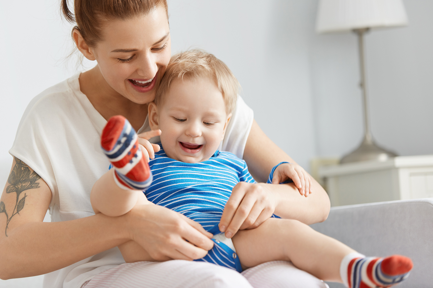 Close-up shot of happy mother and child at home. Young woman buttoning child clothes, carefully holding son on her legs. Cute little boy with blond hair in colorful socks watching her movements. Mother and young child sharing a positive caregiving moment at home.